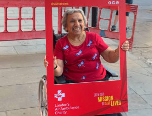 Christies Care client, Gulshan Billimoria, sits smiling in her wheelchair holding a “London’s Air Ambulance Charity” red social media frame board.